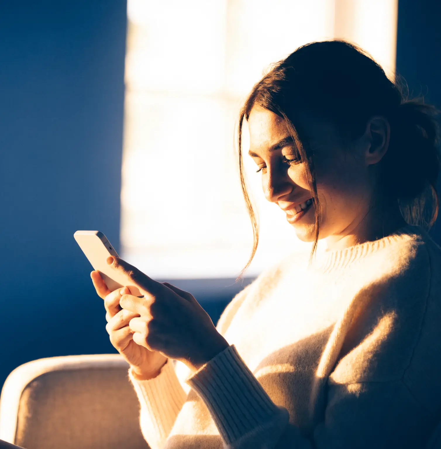 A woman smiles as she reads a reply on her phone in warm morning light.